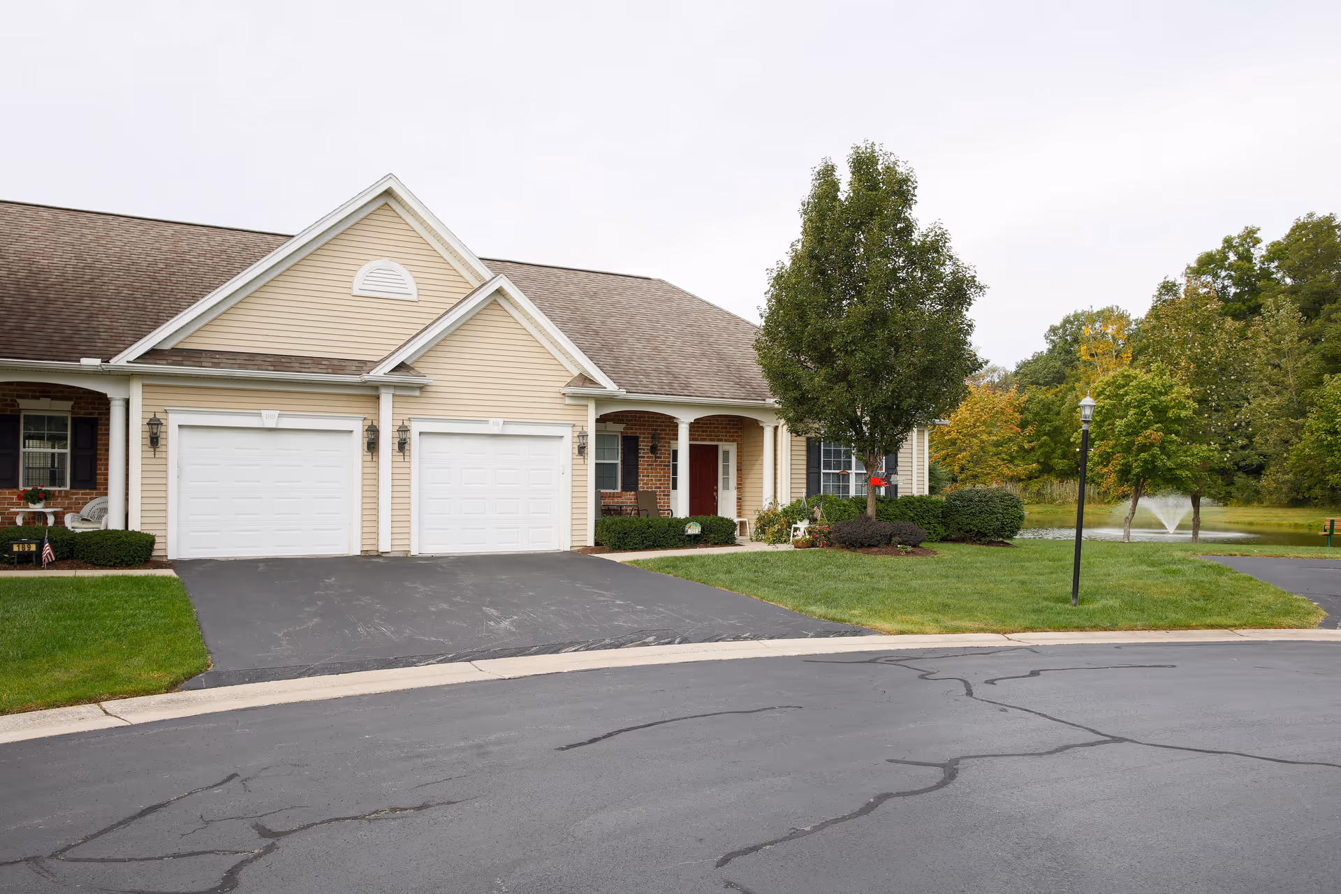 Front exterior of a beige townhome with two garage doors, a driveway, lawn, and trees near a small pond fountain.
