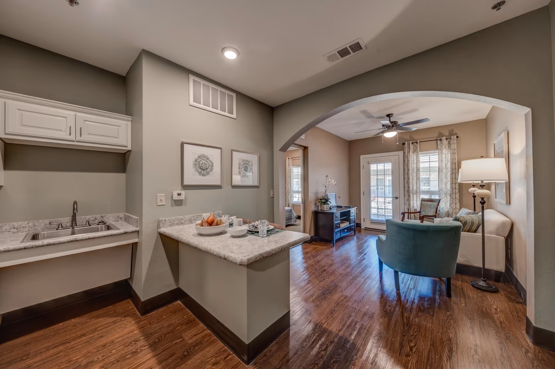 Interior view of an assisted living apartment showing a small kitchen area with a sink and countertop on the left, and a living room with a sofa, armchair, floor lamp, and a ceiling fan. The living room has wooden flooring, a TV stand, and glass doors with curtains leading outside.