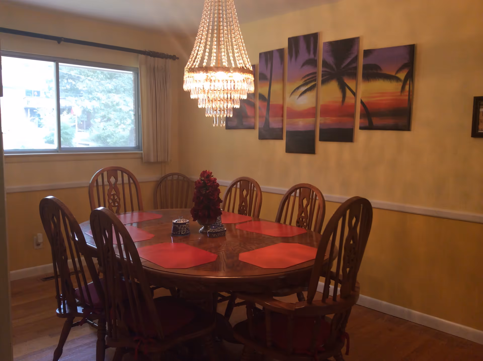 A dining room with a wooden oval table surrounded by eight wooden chairs with red cushions. The table has red placemats and a centerpiece with red flowers. A large crystal chandelier hangs above the table. On the wall, there is a five-panel painting of a sunset with palm trees. A window with beige curtains is on the left side of the room.