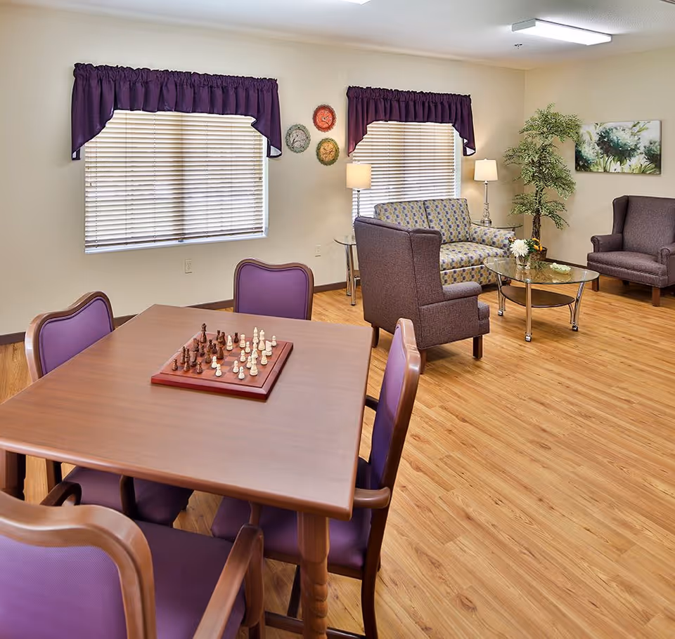 A cozy common room with a wooden table and four purple cushioned chairs arranged around it. A chessboard with pieces set up is on the table. In the background, there are two windows with purple valances, a patterned loveseat, two purple armchairs, a glass coffee table with a small flower arrangement, two floor lamps, a potted plant, and a floral wall painting. The room has light-colored walls and wood flooring.