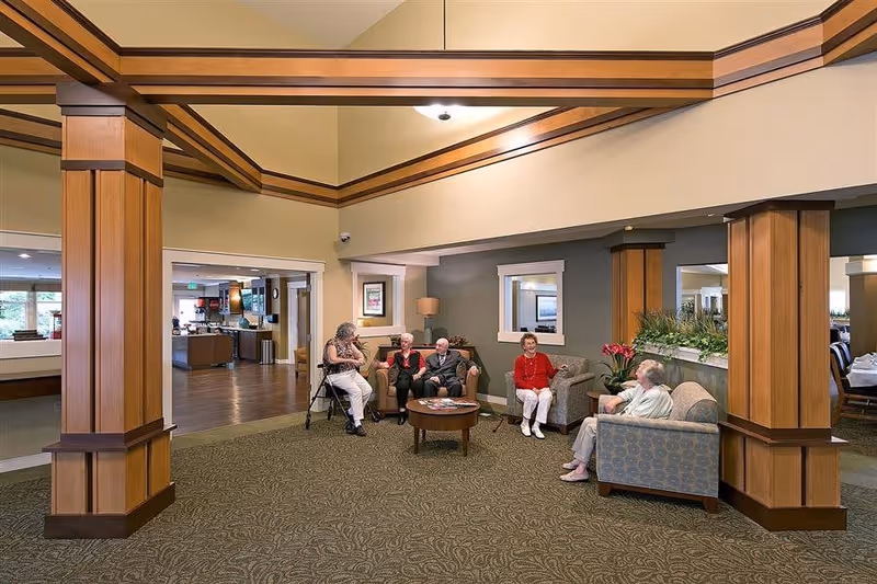 A group of elderly people sitting and chatting in a spacious senior living facility common area with wooden pillars, carpeted floor, and comfortable armchairs. The room has warm lighting and a view into a dining or kitchen area in the background.