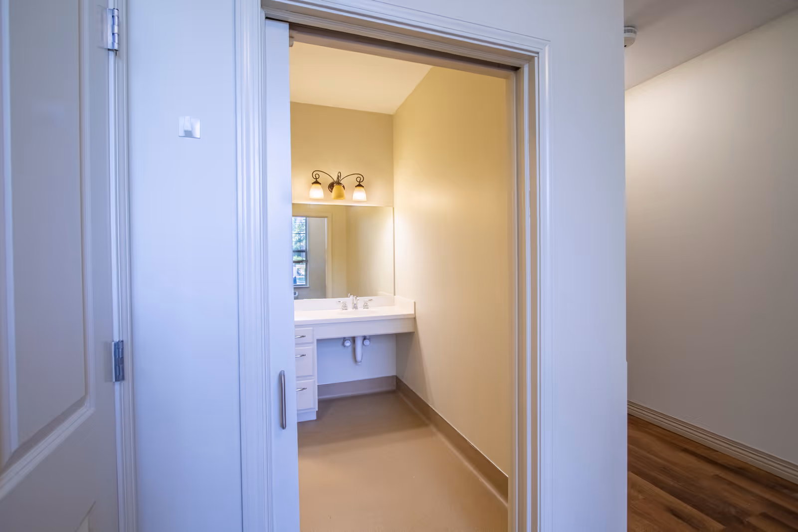 View through a doorway into a small bathroom with a wall-mounted sink, mirror, and a three-light vanity fixture.