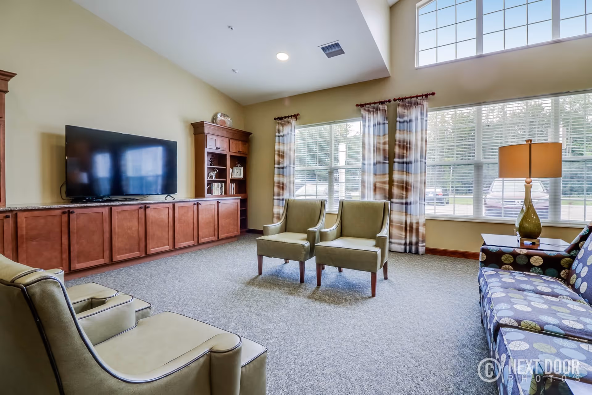 A spacious living room with large windows covered by patterned curtains, allowing natural light to fill the room. The room features a large flat-screen TV mounted on a wooden cabinet with shelves, two green armchairs, a patterned sofa, and a side table with a lamp. The carpeted floor and neutral wall colors create a warm and inviting atmosphere.