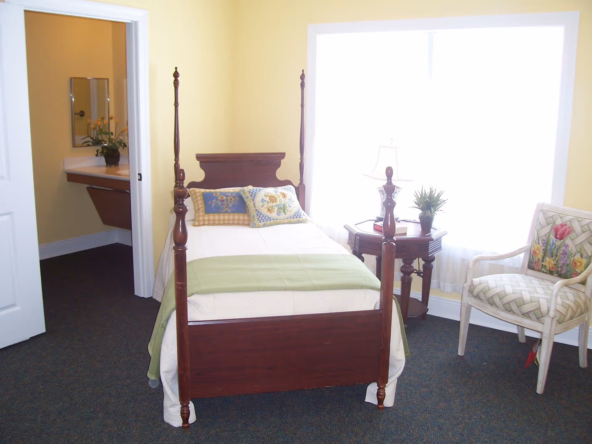 A small bedroom with a wooden four-poster bed made with white bedding, a green throw, and decorative pillows. Next to the bed is a wooden side table with a lamp, a book, and a small plant. There is a cushioned chair with a floral pattern near the window, which is covered with sheer white curtains. An open door reveals a bathroom sink with a mirror and a vase of yellow flowers.
