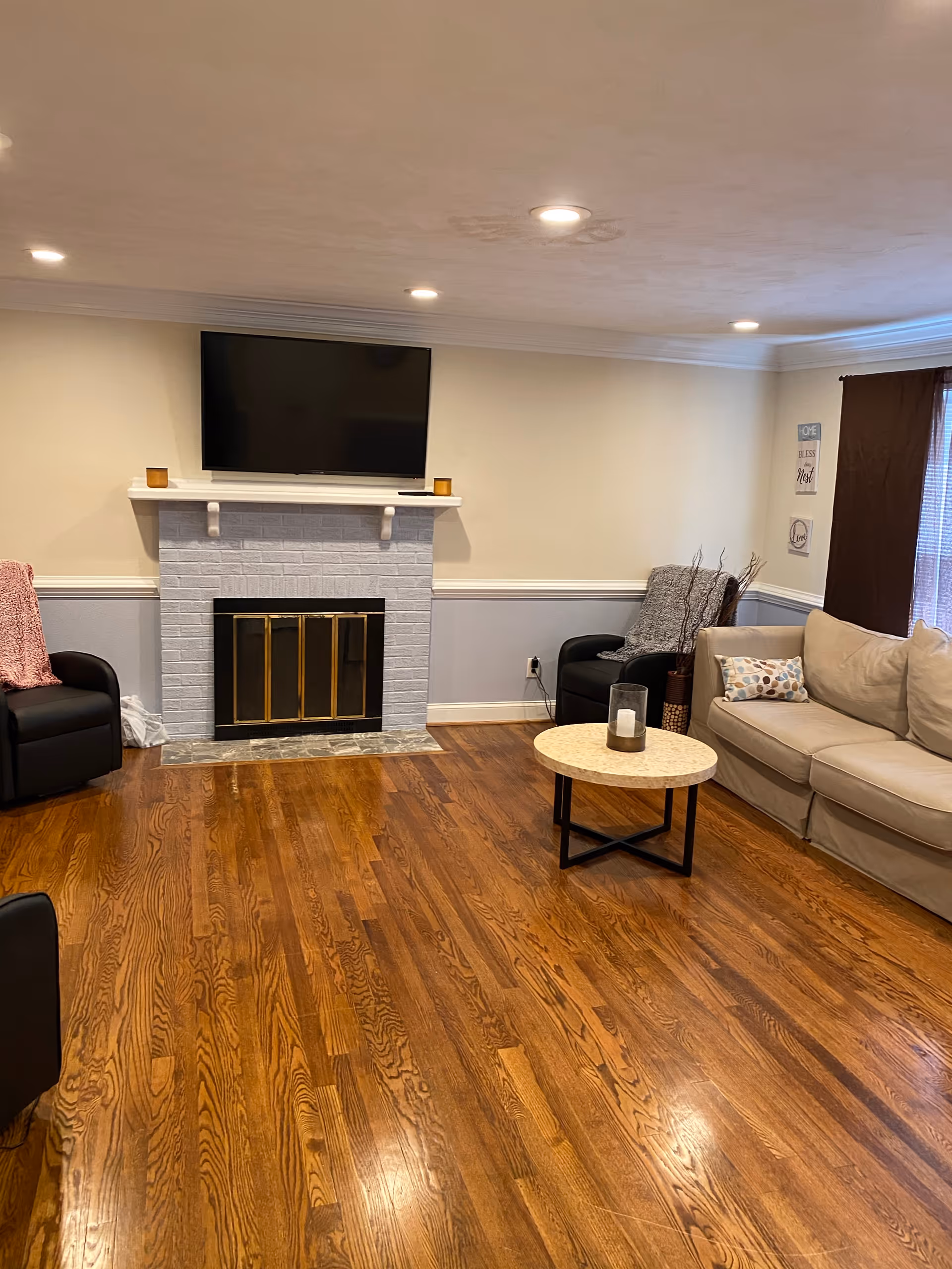 A cozy living room with polished hardwood floors, a white brick fireplace with a mounted flat-screen TV above it, two black armchairs with blankets, a beige sofa with a decorative pillow, a round coffee table with a candle, and a window with brown curtains.