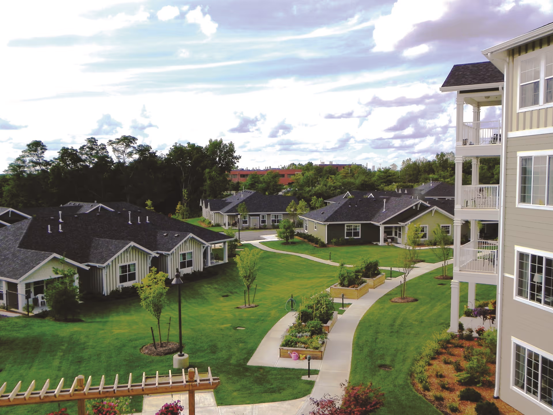 Courtyard view of a senior living community with walking paths, raised garden beds, single-story cottages, and a multi-story building with balconies.