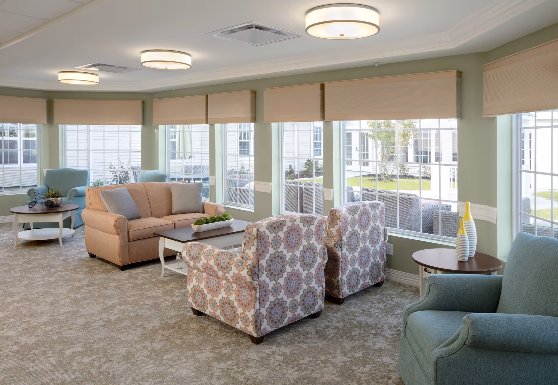 A bright and spacious living room area in an assisted living facility featuring large windows with beige blinds, a beige sofa with two gray cushions, two patterned armchairs, a blue armchair, and two round wooden side tables with decorative items. The room has light green walls, beige carpet, and modern ceiling lights.