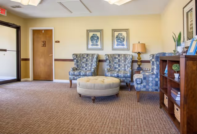 A cozy seating area in a senior living facility with three patterned armchairs arranged around a round beige ottoman. There is a wooden bookshelf with decorative items and books on the right side, and two framed pictures of blue vases on the wall behind the chairs. A table lamp is placed between two of the chairs. The room has beige walls, carpeted floor, and a wooden door with an exit sign above it on the left side.