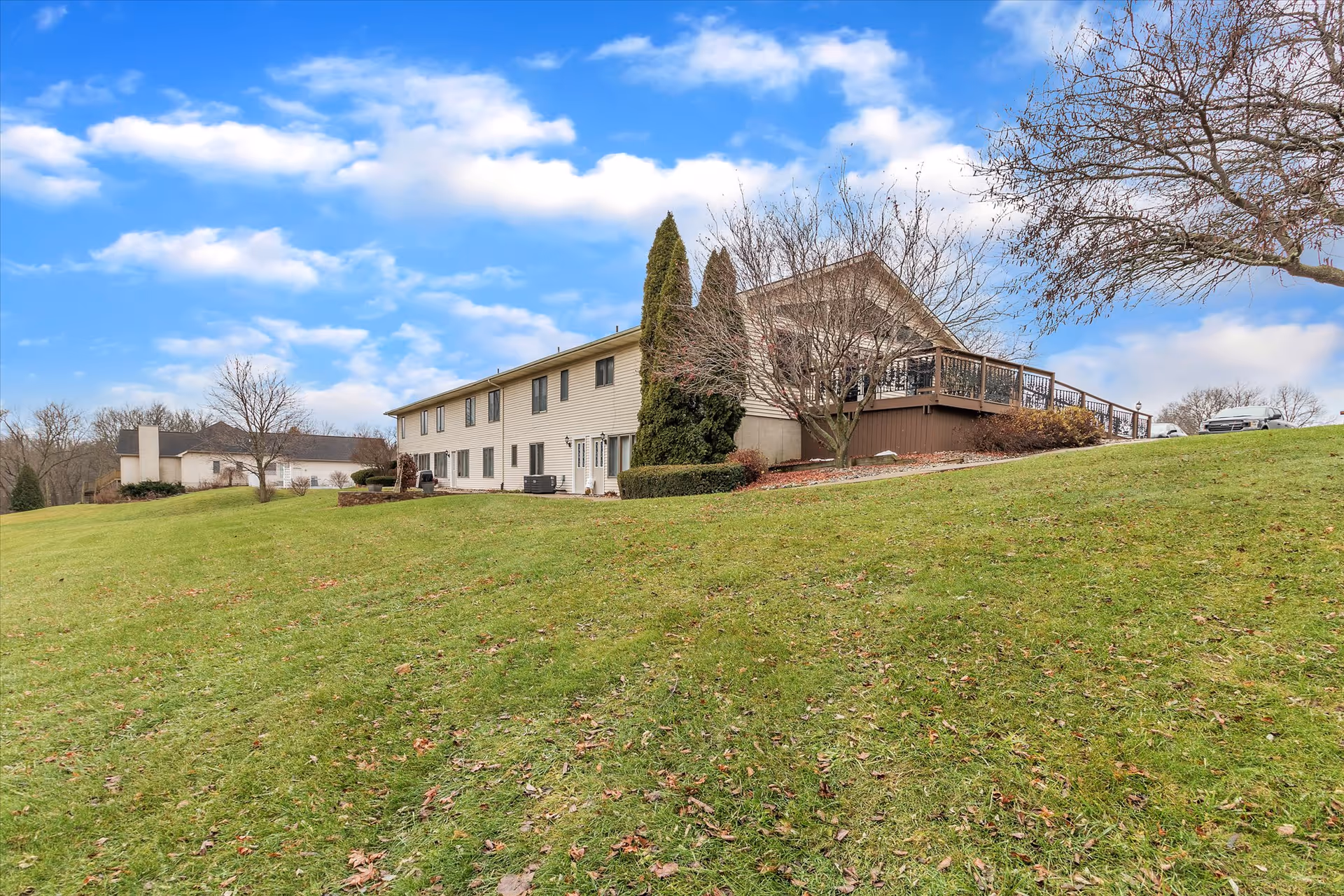 Exterior view of a two-story building with beige siding situated on a grassy hill under a partly cloudy blue sky. The building has multiple windows and a large wooden deck on one side. Leafless trees and some shrubs are visible around the building.
