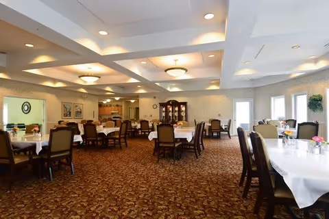 A spacious dining room with multiple tables covered in white tablecloths, each surrounded by wooden chairs with cushioned seats. The room has patterned carpet flooring, soft overhead lighting, and large windows letting in natural light. There is a china cabinet against the far wall and floral centerpieces on the tables.