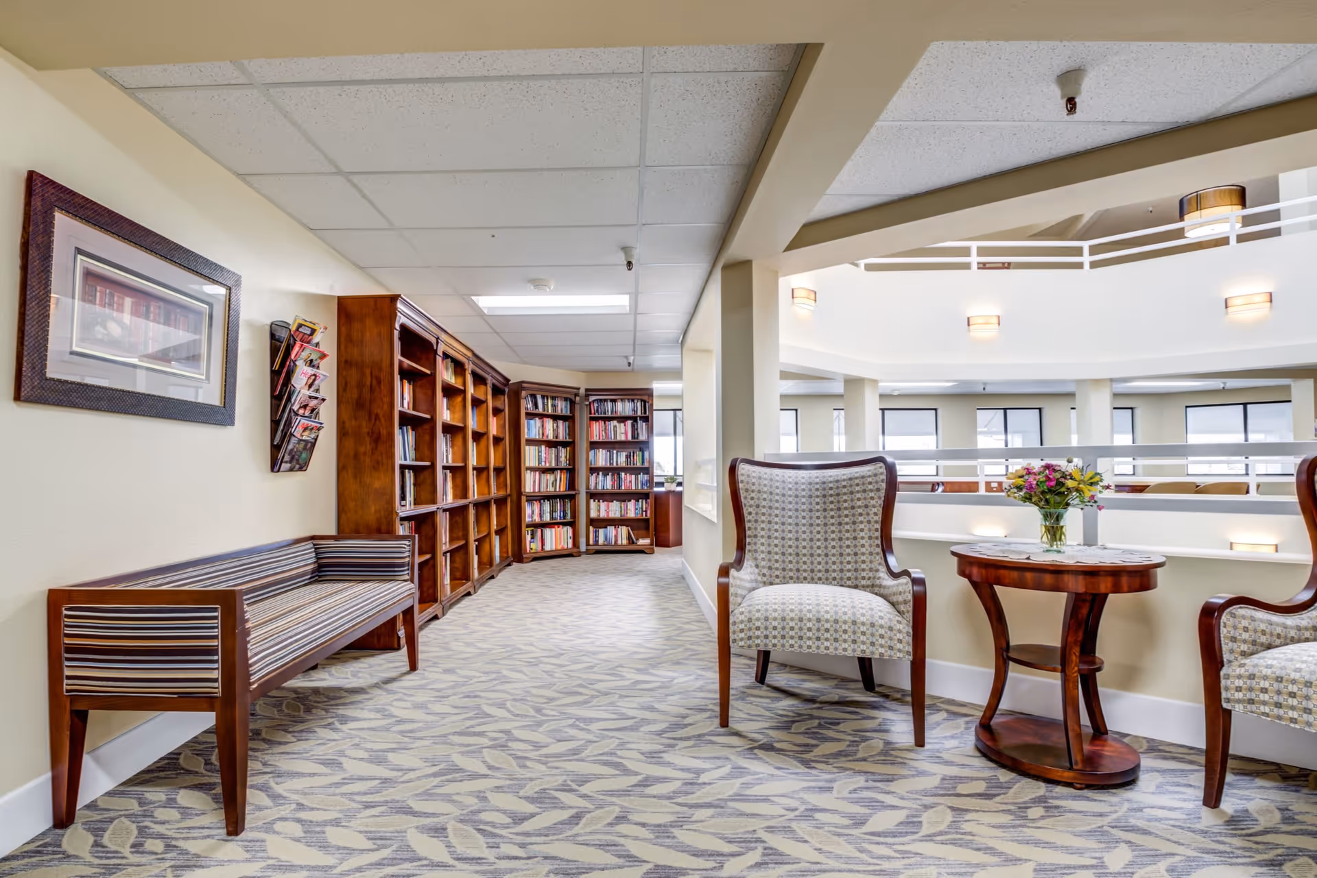 Interior view of a senior living community library area with wooden bookshelves filled with books, a striped wooden bench along the left wall, two upholstered armchairs, and a small wooden table with a vase of flowers. The space overlooks a bright atrium with railings and multiple windows.