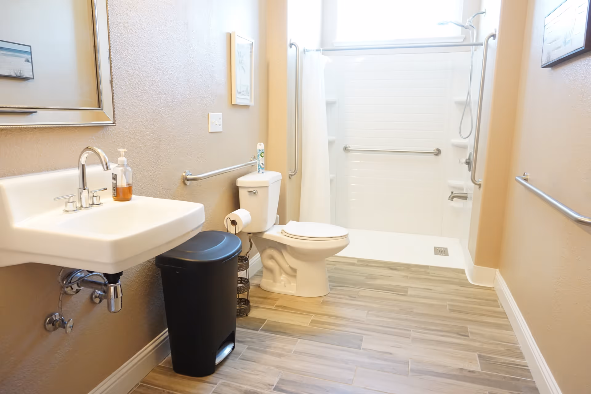 A clean and accessible bathroom with a white sink, a black trash bin, a toilet, and a walk-in shower with grab bars and a shower curtain. The floor has wood-like tiles and the walls are painted beige with framed pictures hanging.