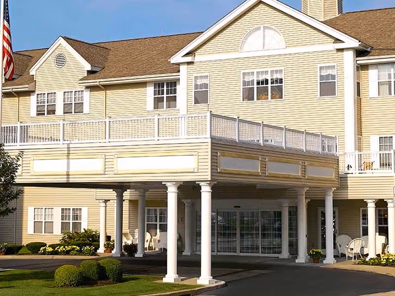 Exterior view of a senior living facility building with beige siding, white columns supporting a large balcony, multiple windows, and an American flag on the left side. There are shrubs and a paved driveway in front of the entrance.