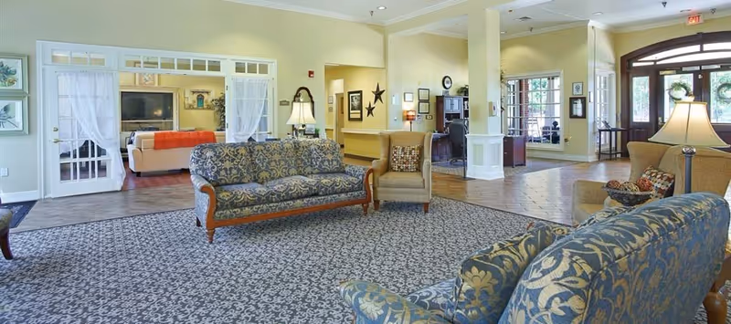 Bright, furnished senior living lounge with patterned sofas, armchairs, lamps, and a view toward a reception area and entry doors.