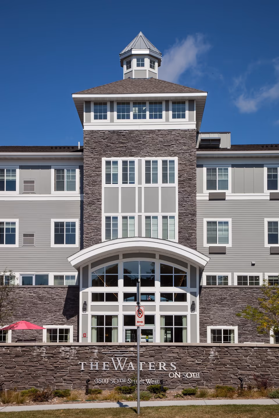 Front exterior view of The Waters on 50th senior living facility, featuring a multi-story building with gray stone and siding, large windows, and a prominent entrance with an arched white frame. A stone wall in front displays the facility name and address. The sky is clear and blue.