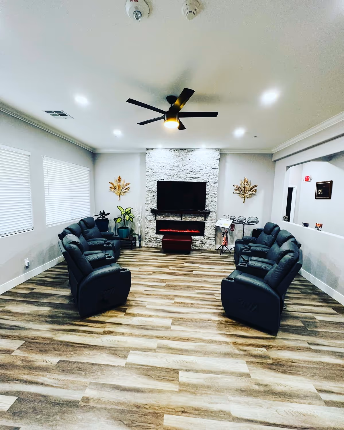 A modern living room with two rows of black recliner chairs facing a wall-mounted flat-screen TV above an electric fireplace. The room has wood-look flooring, white walls, two windows with blinds on the left, decorative wall art, potted plants, and a ceiling fan with lights.