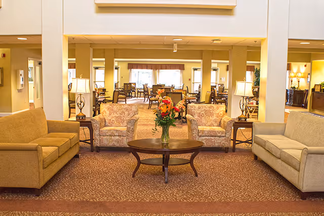 A spacious senior living facility common area with two beige sofas and two patterned armchairs arranged around a wooden coffee table with a vase of flowers. The room has warm lighting from table lamps and a carpeted floor, with a dining area visible in the background.