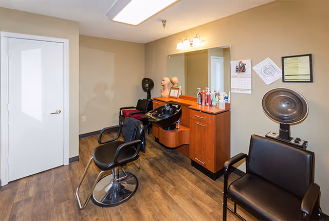 Interior view of a hair salon area in a senior living facility with a black salon chair in front of a hair washing station, a wooden cabinet with various hair care products on top, a large mirror, two mannequin heads, a hair dryer chair, and a closed white door.