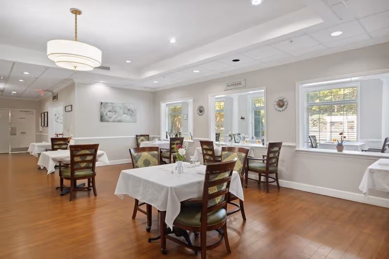 A bright dining room with wooden floors and several tables covered with white tablecloths. Each table is surrounded by wooden chairs with green cushioned seats and floral-patterned backs. The walls are painted light gray with white trim, and there are three large windows letting in natural light. Decorative wall art and small plants are placed on the windowsills. A modern circular ceiling light fixture hangs above the room.