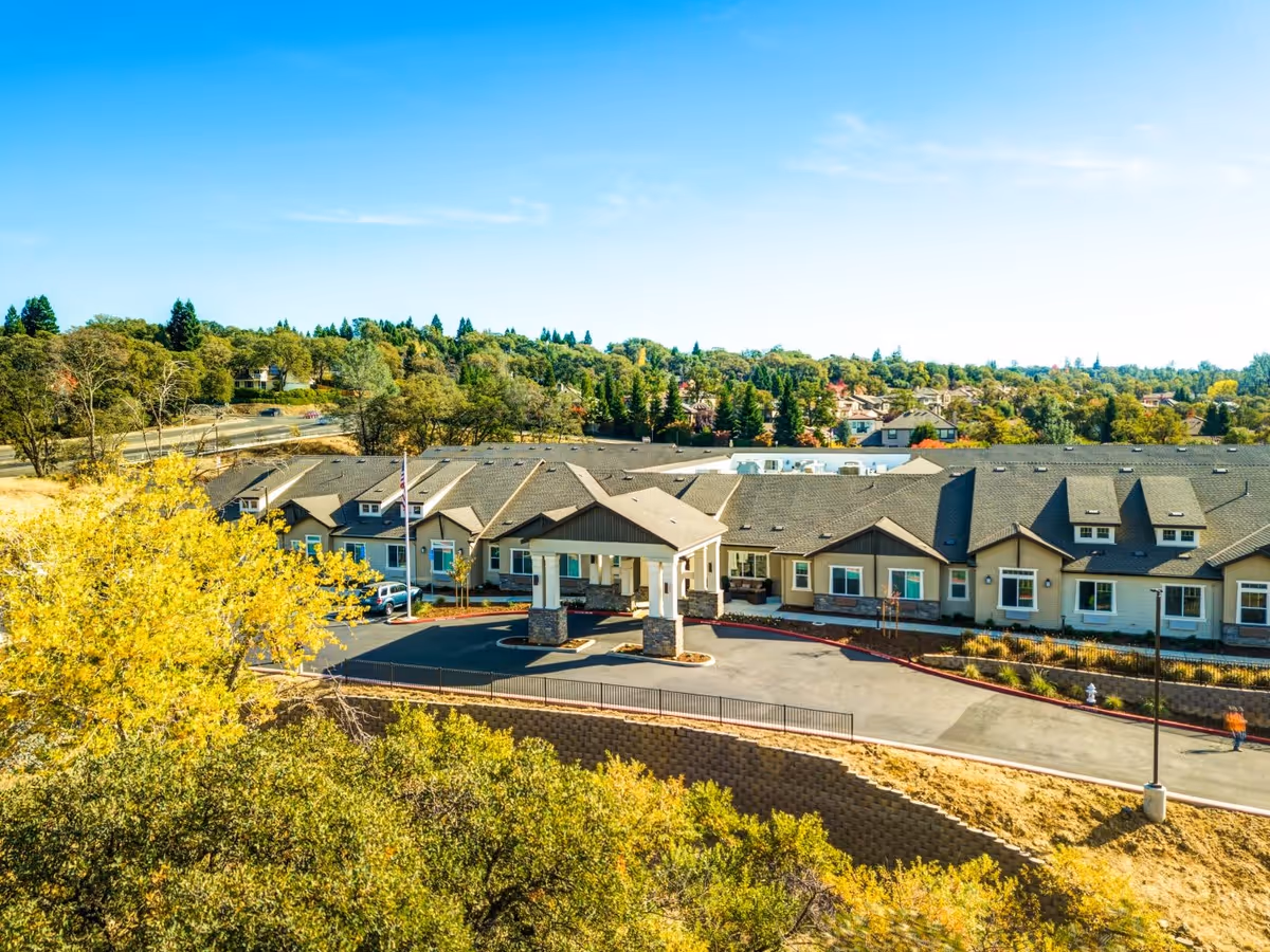 Aerial view of The Pavilion at El Dorado Hills senior living facility showing a large single-story building with a covered entrance, surrounded by trees and greenery under a clear blue sky.