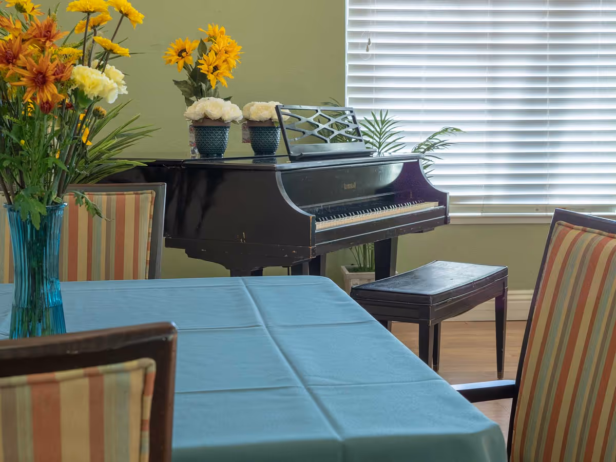 Indoor common room with a black piano by a window, a table covered with a blue tablecloth and a vase of flowers, and striped dining chairs.