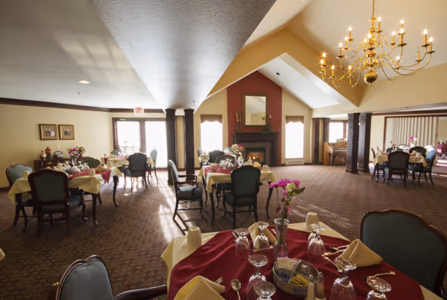 A spacious dining room with multiple round tables covered with red and yellow tablecloths, set with glasses, napkins, and silverware. The room features a high vaulted ceiling with a large chandelier, a fireplace with a mirror above it, and several windows letting in natural light. The carpet is patterned, and there are upholstered chairs around the tables.