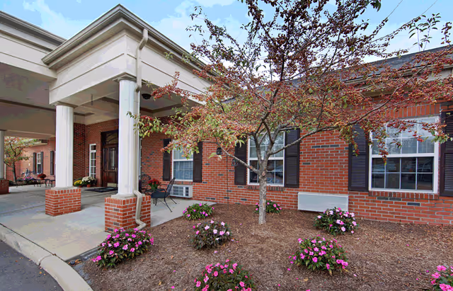 Exterior view of a senior living facility showing a brick building with white columns supporting a covered entrance. There are windows with black shutters, a small tree with red leaves, and flower beds with pink flowers in front of the building.