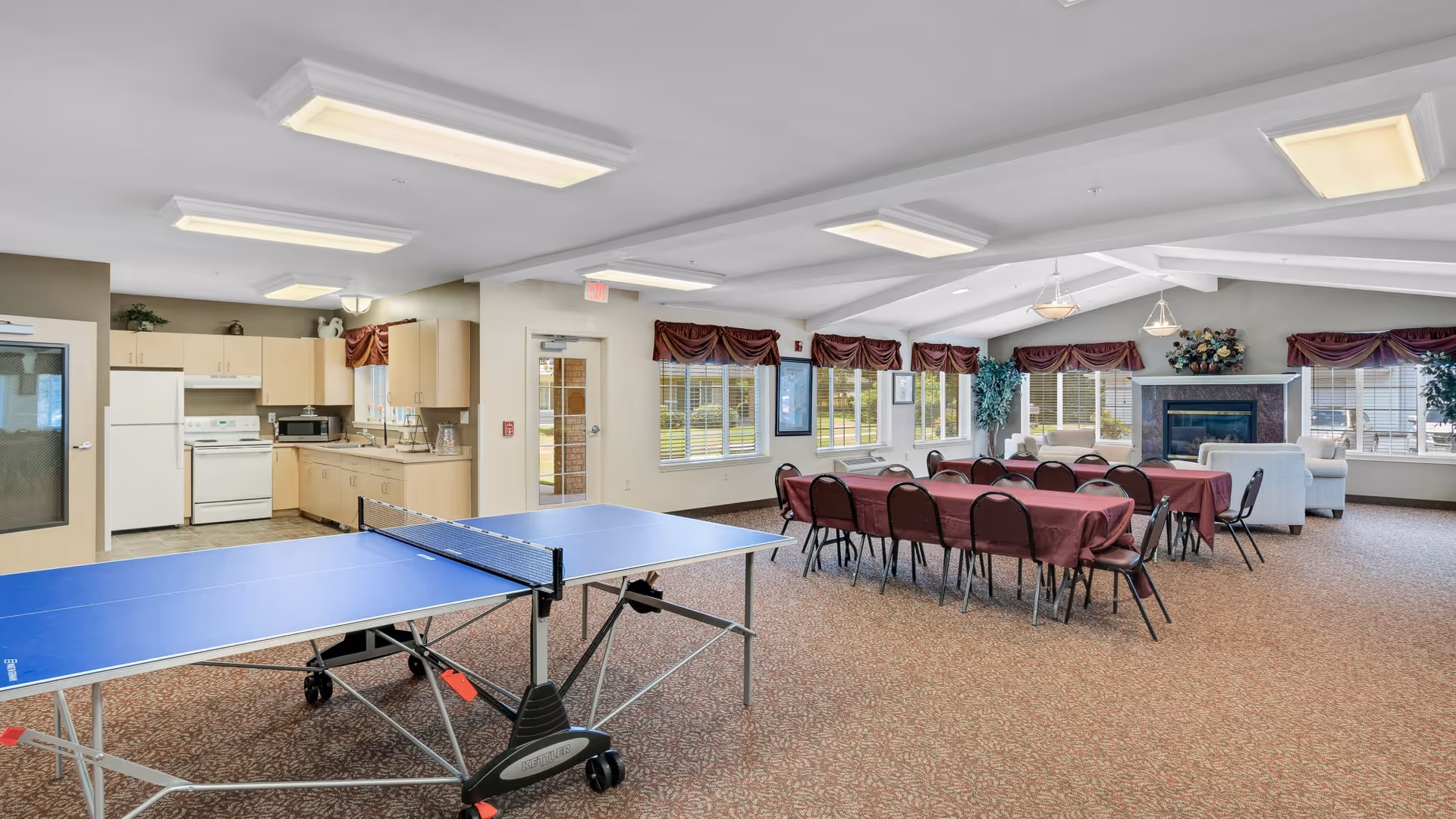 A spacious common area in a senior living facility featuring a blue ping pong table in the foreground, a kitchen with light wood cabinets and white appliances on the left, and a dining area with tables covered in maroon tablecloths and chairs. The room has large windows with maroon valances, carpeted floors, and a seating area with sofas near a fireplace decorated with a floral arrangement.