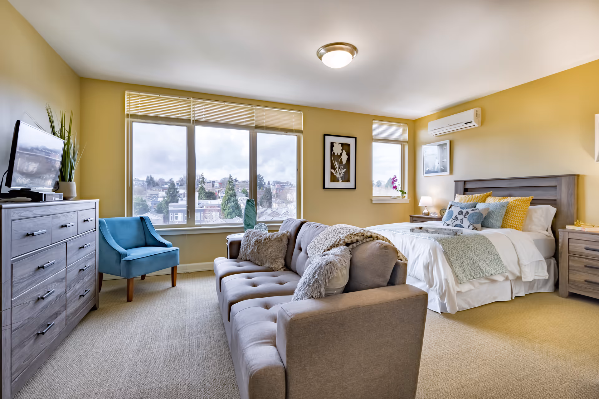 A bright and spacious bedroom with a large window showing an outside view. The room features a bed with multiple pillows and a light blanket, a gray sofa with throw pillows, a blue armchair, a wooden dresser with a TV on top, and a nightstand with a lamp. The walls are painted yellow and there is a framed floral artwork on the wall.