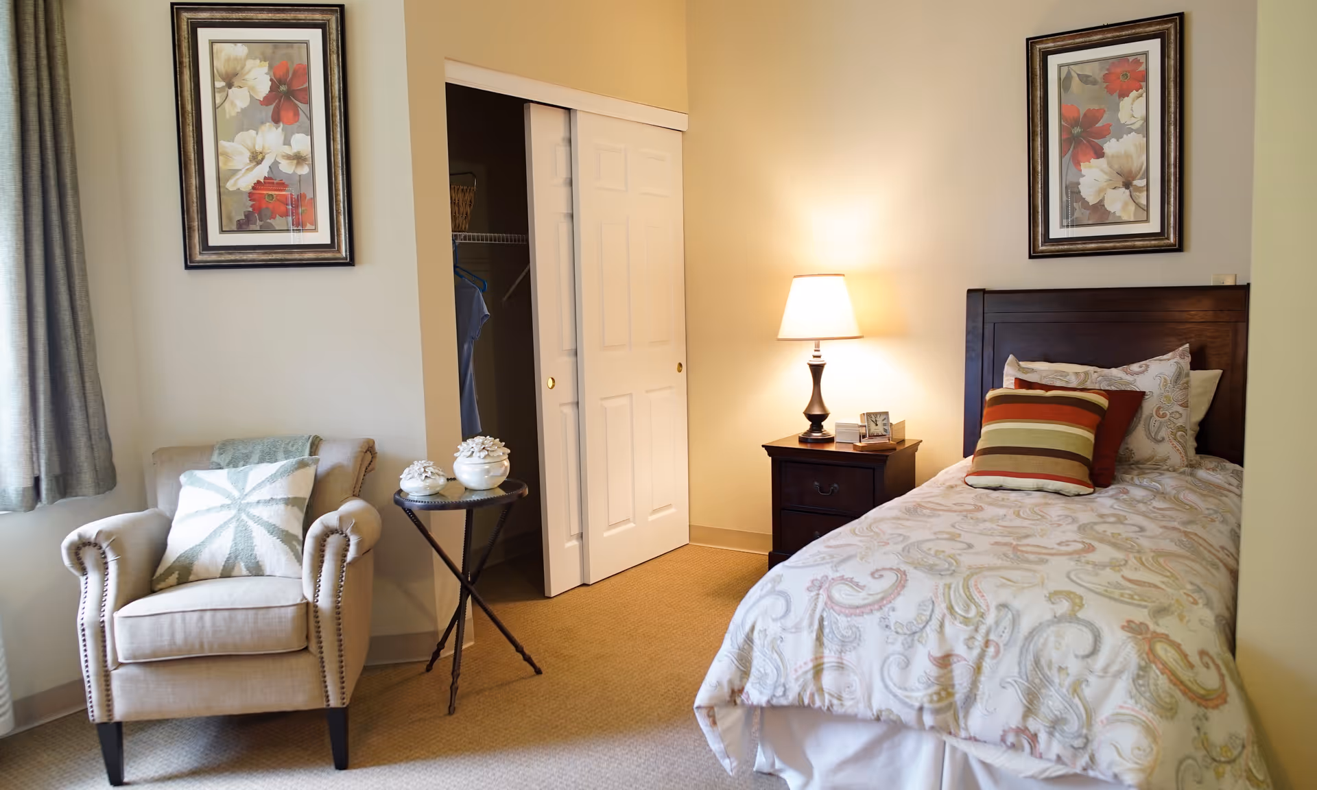 Neatly furnished bedroom with a single bed, nightstand and lamp, armchair, small side table and an open closet.