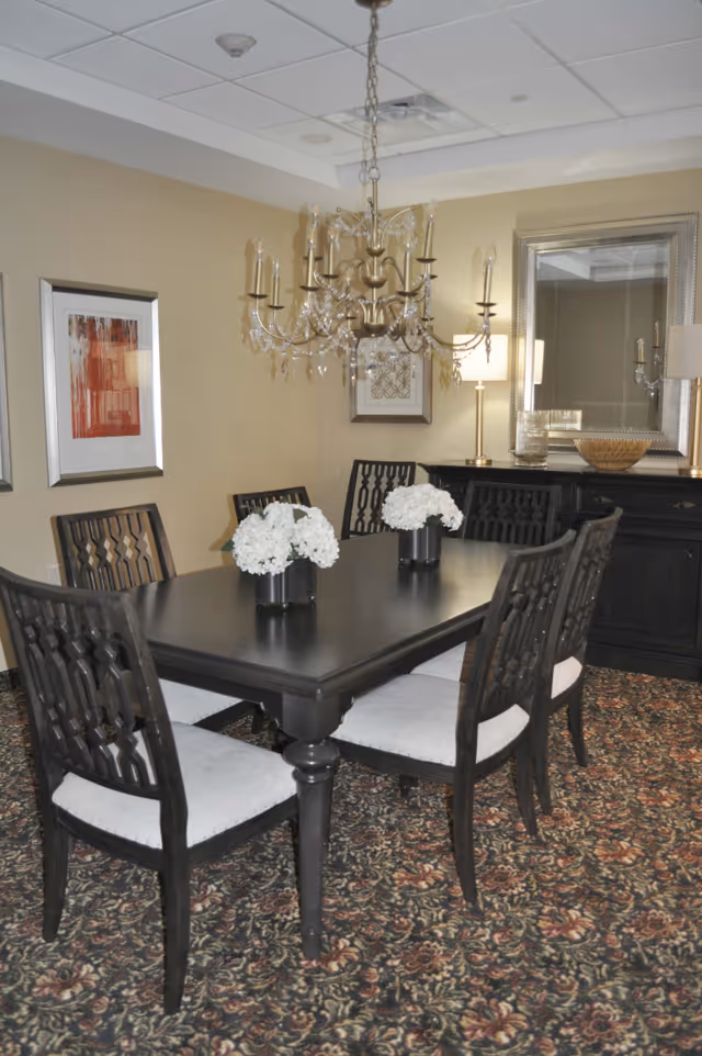 A formal dining room with a dark wooden table and six matching chairs with light-colored cushions. The table is decorated with two small vases of white flowers. Above the table hangs an ornate chandelier with multiple candle-style lights and crystal accents. The room has beige walls adorned with framed artwork, a large mirror, and a black sideboard with lamps and decorative items. The floor is covered with a patterned carpet.