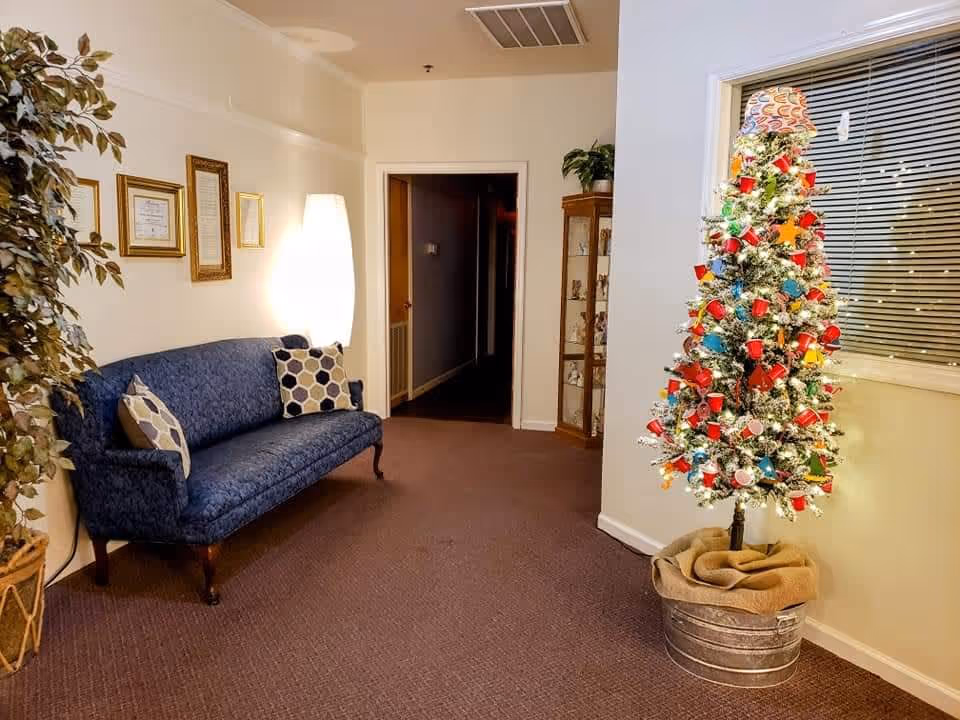 Interior hallway of a senior living facility with a blue upholstered couch with patterned pillows on the left, a tall white floor lamp behind it, framed certificates on the wall, a potted plant, and a small decorated Christmas tree in a metal bucket on the right near a window with blinds.