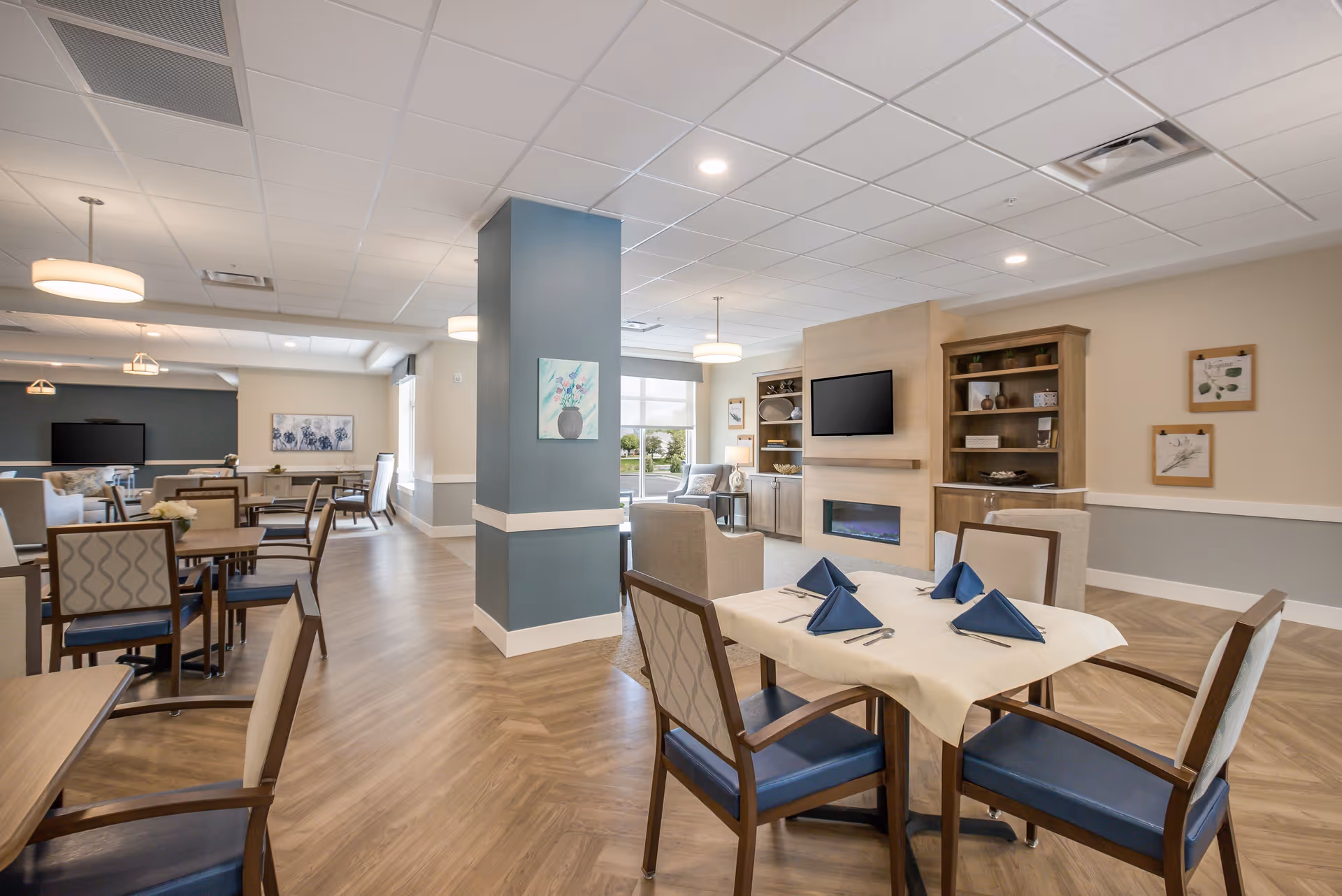 Bright communal dining room with set tables and chairs and a lounge area featuring a fireplace and TV.