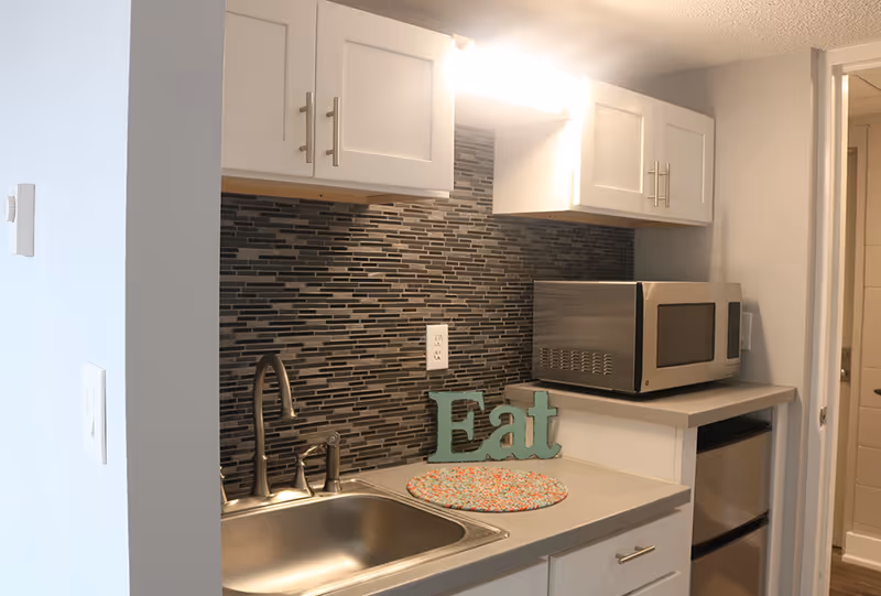 Small kitchen area with a stainless steel sink, modern faucet, mosaic tile backsplash, white cabinets, a microwave on the counter, a small refrigerator below, and a decorative sign that says 'Eat'.