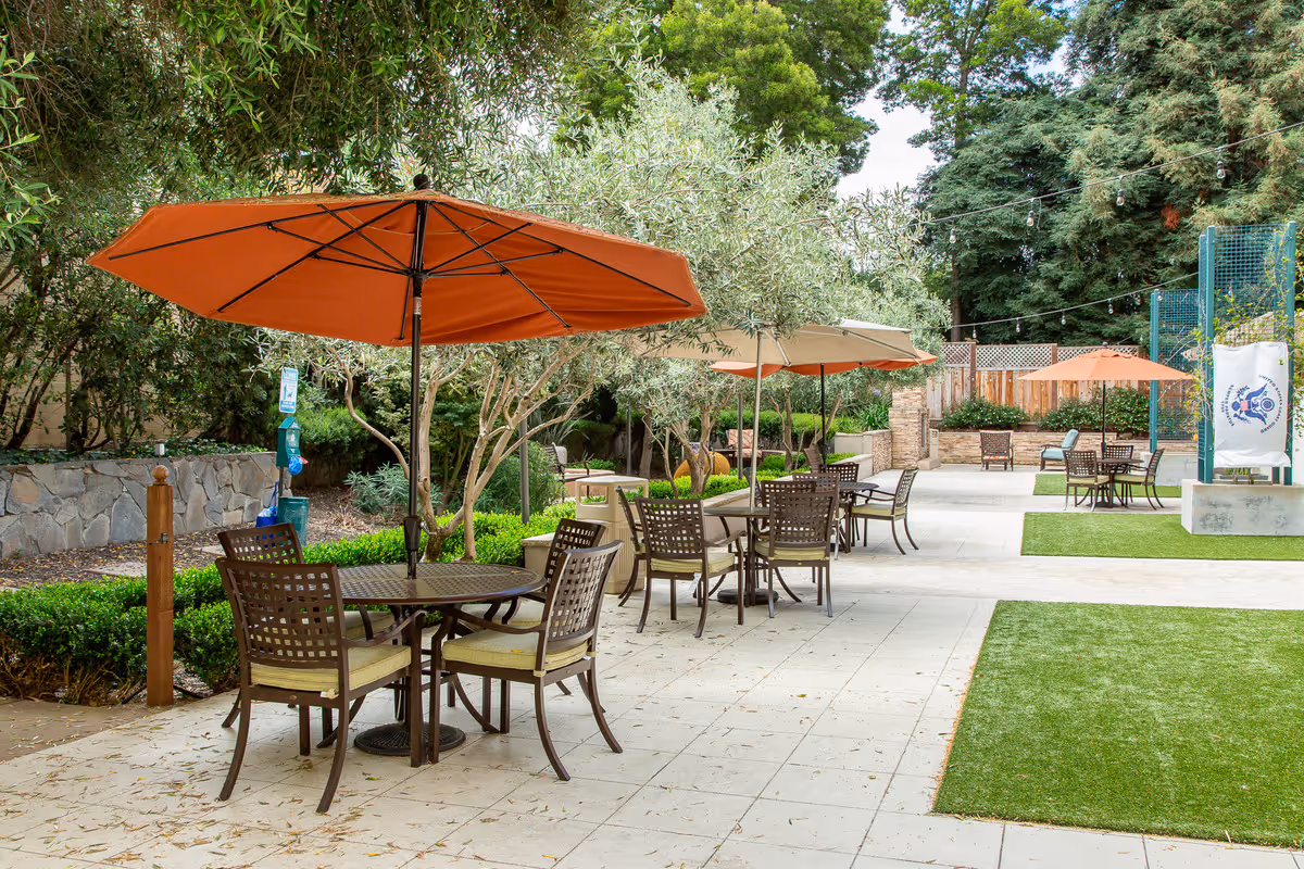 Outdoor patio area with multiple round tables and chairs, each shaded by large orange and beige umbrellas. The space is surrounded by greenery including trees and bushes, with string lights overhead and a stone wall on one side.