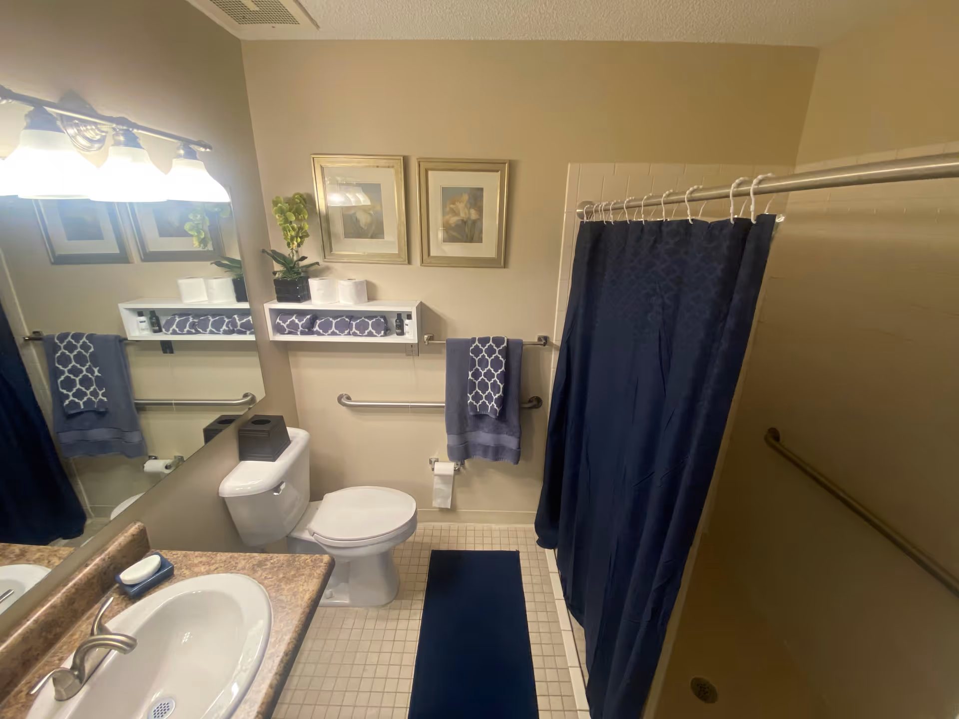 A clean bathroom with a white toilet, a sink with a marble countertop, a large mirror, and a shower with a dark blue curtain. There are two framed floral pictures on the wall above a small white shelf holding a plant, toilet paper rolls, and toiletries. A dark blue towel hangs on a grab bar next to the toilet, and a matching dark blue bath mat is on the tiled floor.