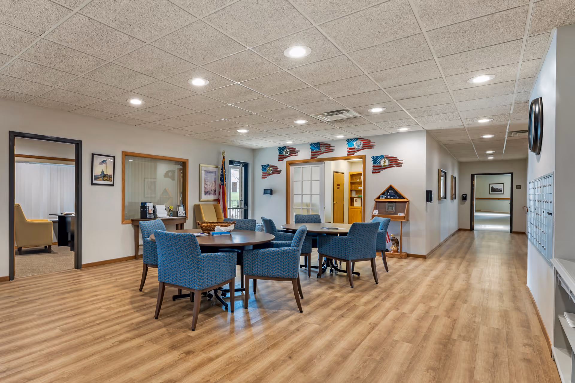 A spacious, well-lit common area in a senior living facility with wooden flooring, round tables surrounded by blue upholstered chairs, and a hallway leading to other rooms. The walls are decorated with framed pictures, American flag-themed wall art, and a display case with a folded flag. There is a door with glass panels and an American flag standing in the corner.