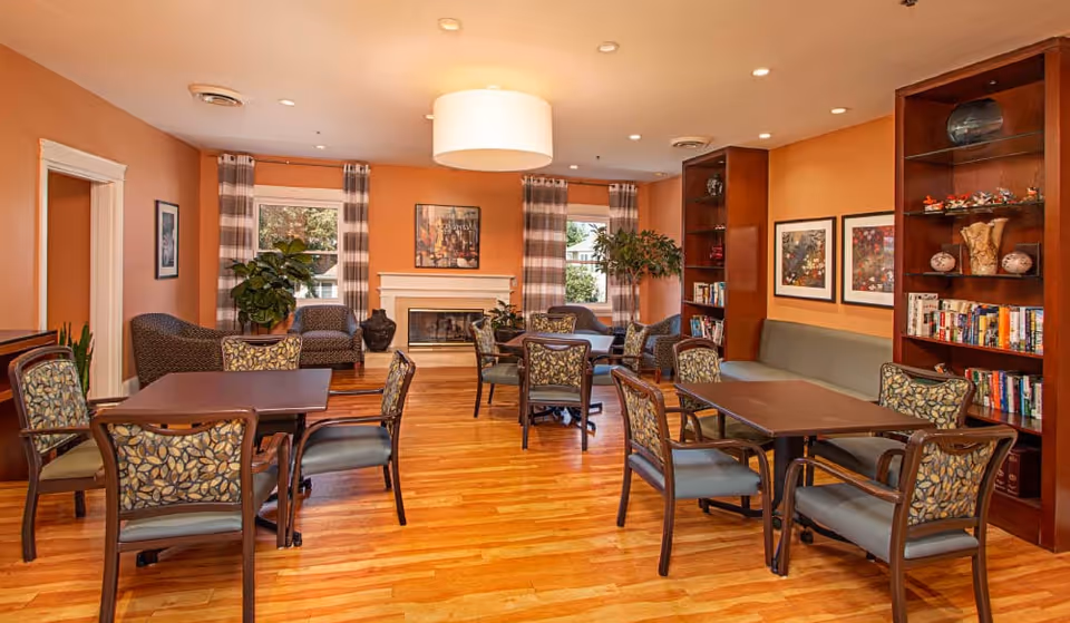 Bright communal lounge with wooden floors, multiple tables and upholstered chairs, bookshelves and a fireplace.
