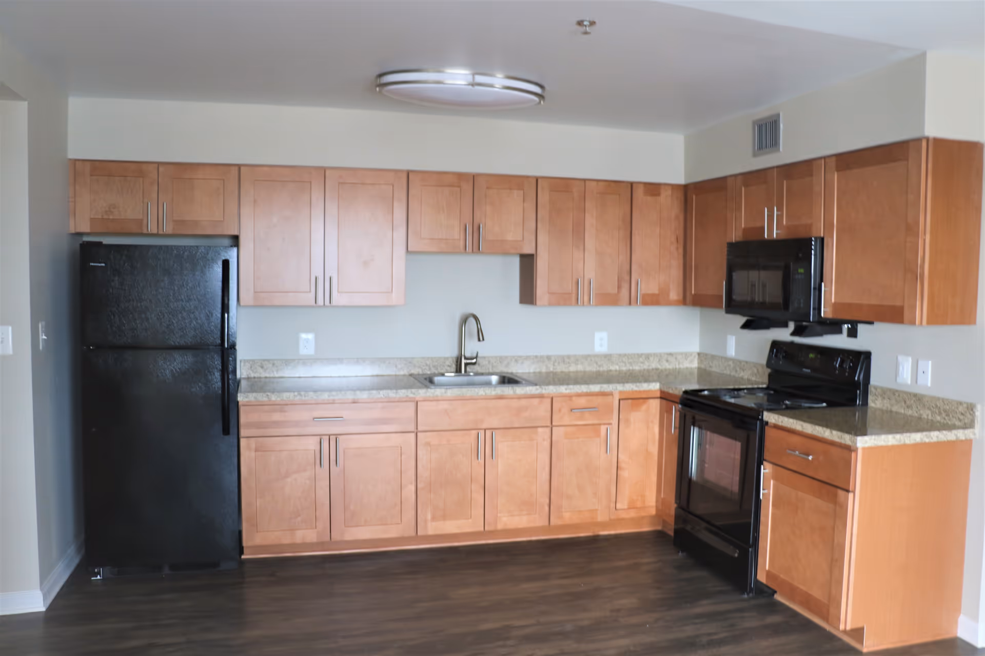 A modern kitchen with light brown wooden cabinets, a black refrigerator, a black stove with an oven, a black microwave mounted above the stove, a stainless steel sink with a faucet, and granite countertops. The floor is dark wood, and the walls are light-colored.