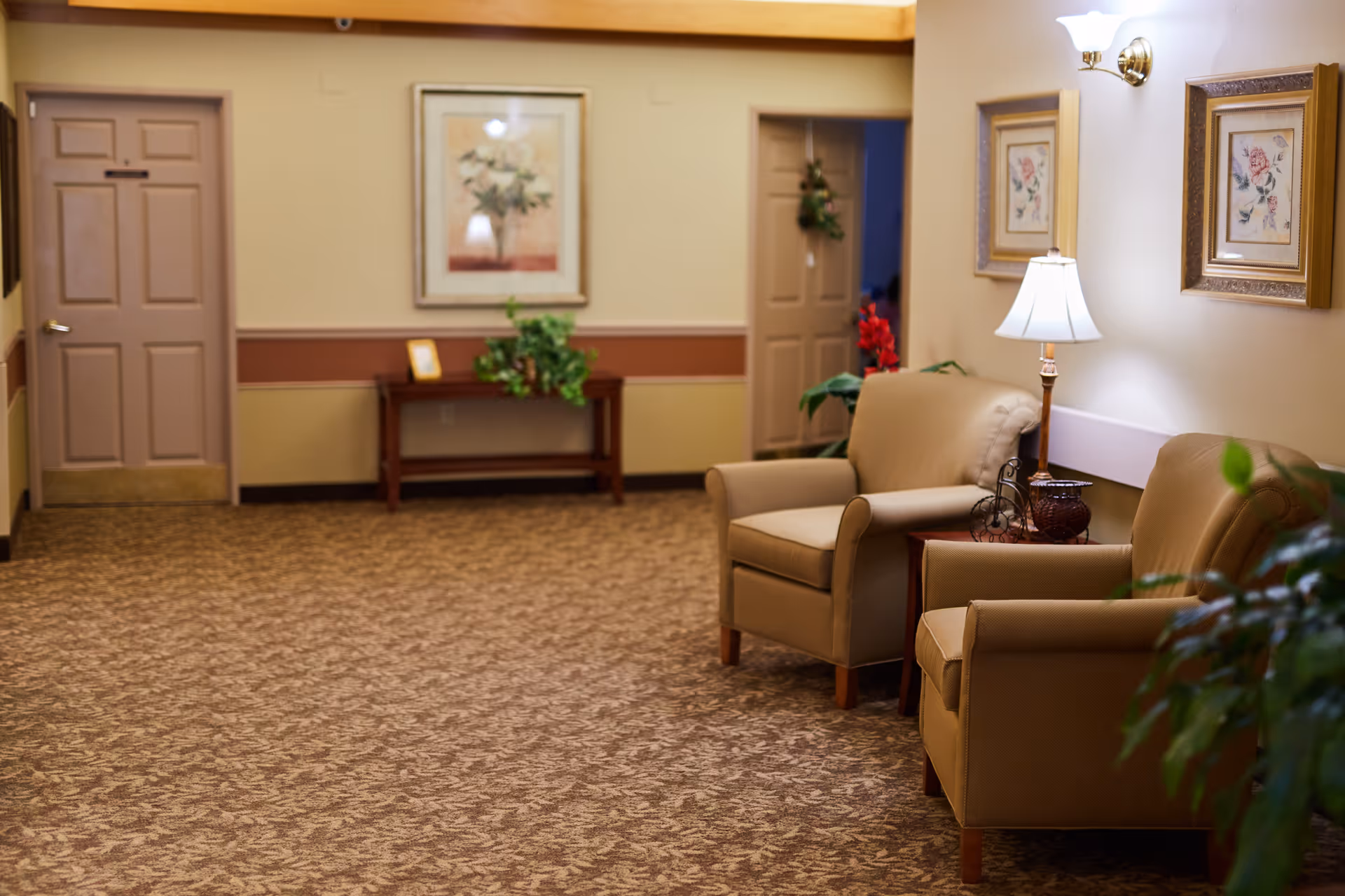 A cozy senior living facility hallway with two beige armchairs, a small wooden side table with a lamp and decorative items, framed floral artwork on the walls, a patterned carpet, and two closed doors. There is also a wooden console table with a plant and a picture frame against the far wall.