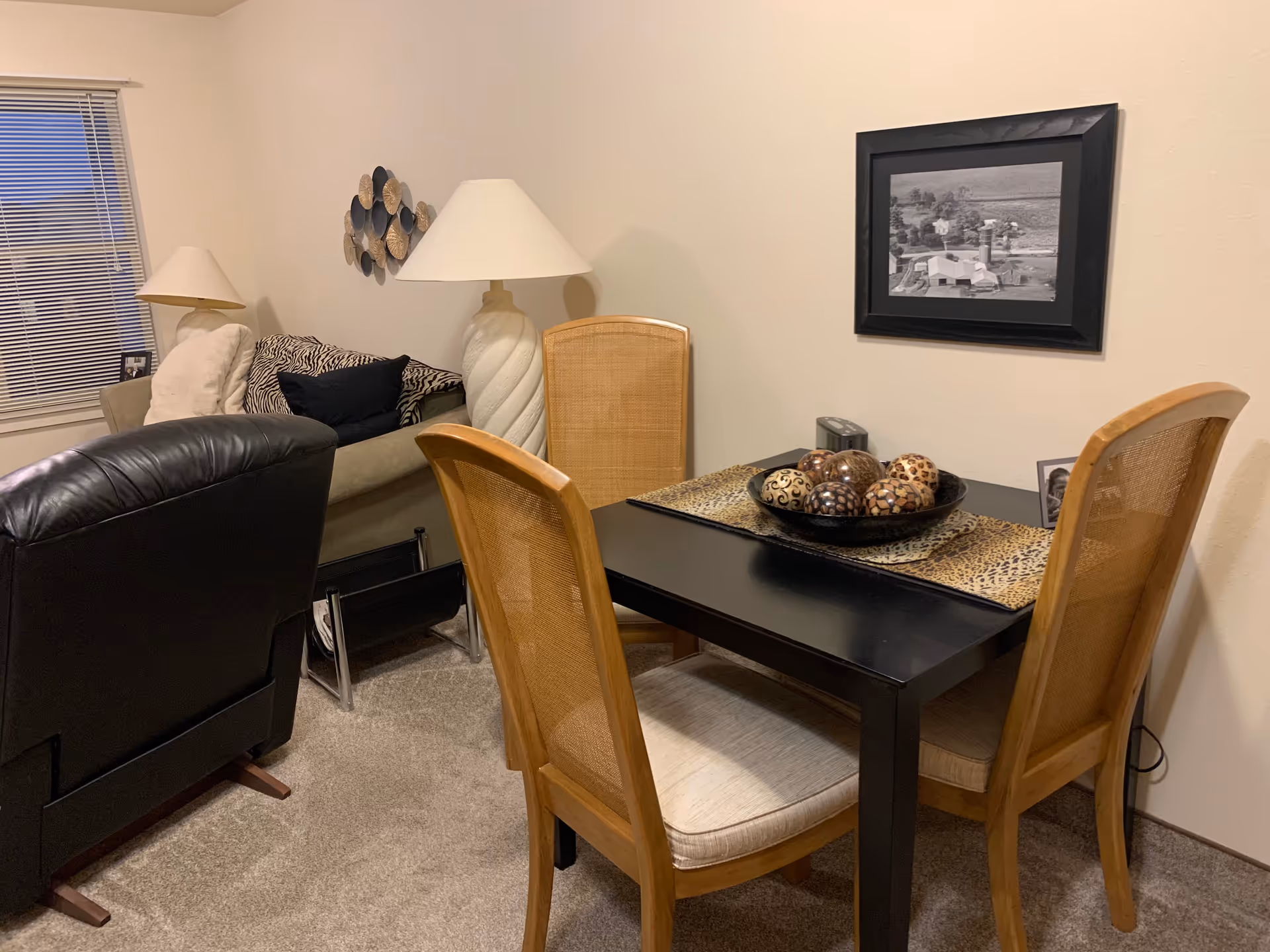 A cozy living and dining area with a black dining table surrounded by four wooden chairs with cushioned seats. On the table is a decorative bowl filled with patterned spheres and a leopard print table runner. Behind the table is a beige wall with a black and white framed photograph. To the left, there is a beige couch with patterned pillows and a white throw blanket, next to a large white lamp. A black leather recliner is partially visible in the foreground, and a window with closed blinds is on the far left wall.