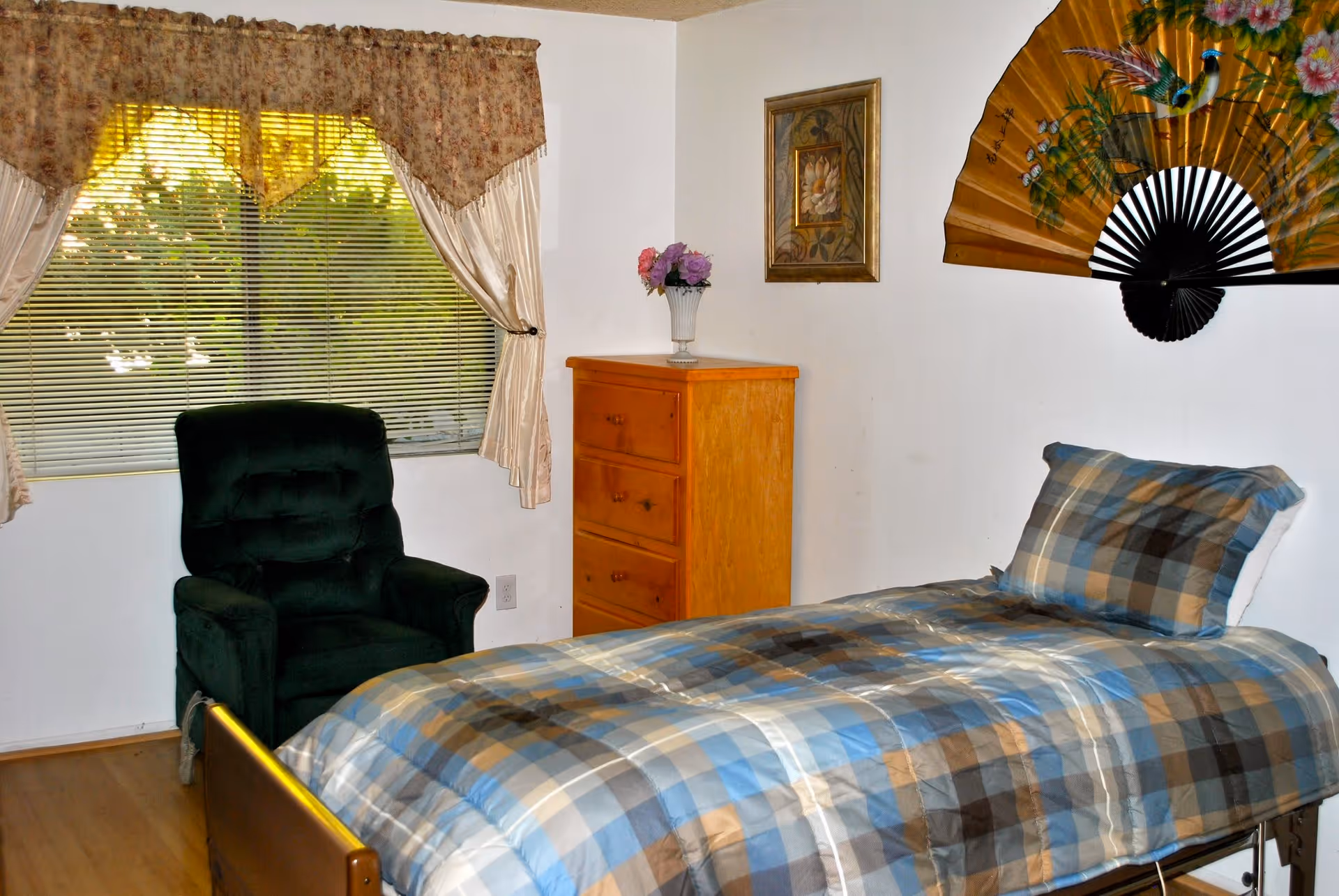 A bedroom with a single bed covered in a blue and brown plaid comforter and matching pillow. Next to the bed is a wooden dresser with a vase of flowers on top. A dark green armchair is positioned near a window with floral curtains and blinds. On the wall above the bed is a decorative fan and a framed picture.