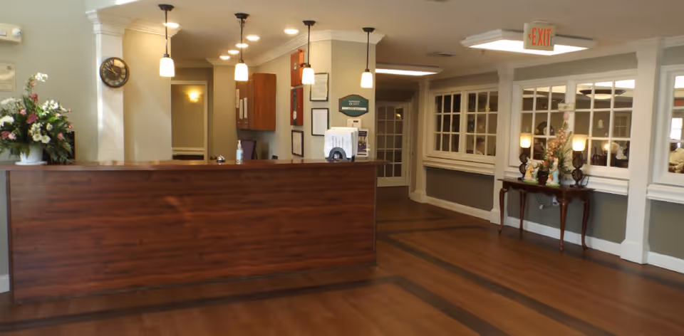 Reception area of Pee Dee Gardens facility featuring a wooden front desk with hanging pendant lights above, a flower arrangement on the left side, a clock on the wall, and a hallway leading to other rooms. There is a small table with decorative items and lamps against the right wall under large windows, and an exit sign above a doorway.