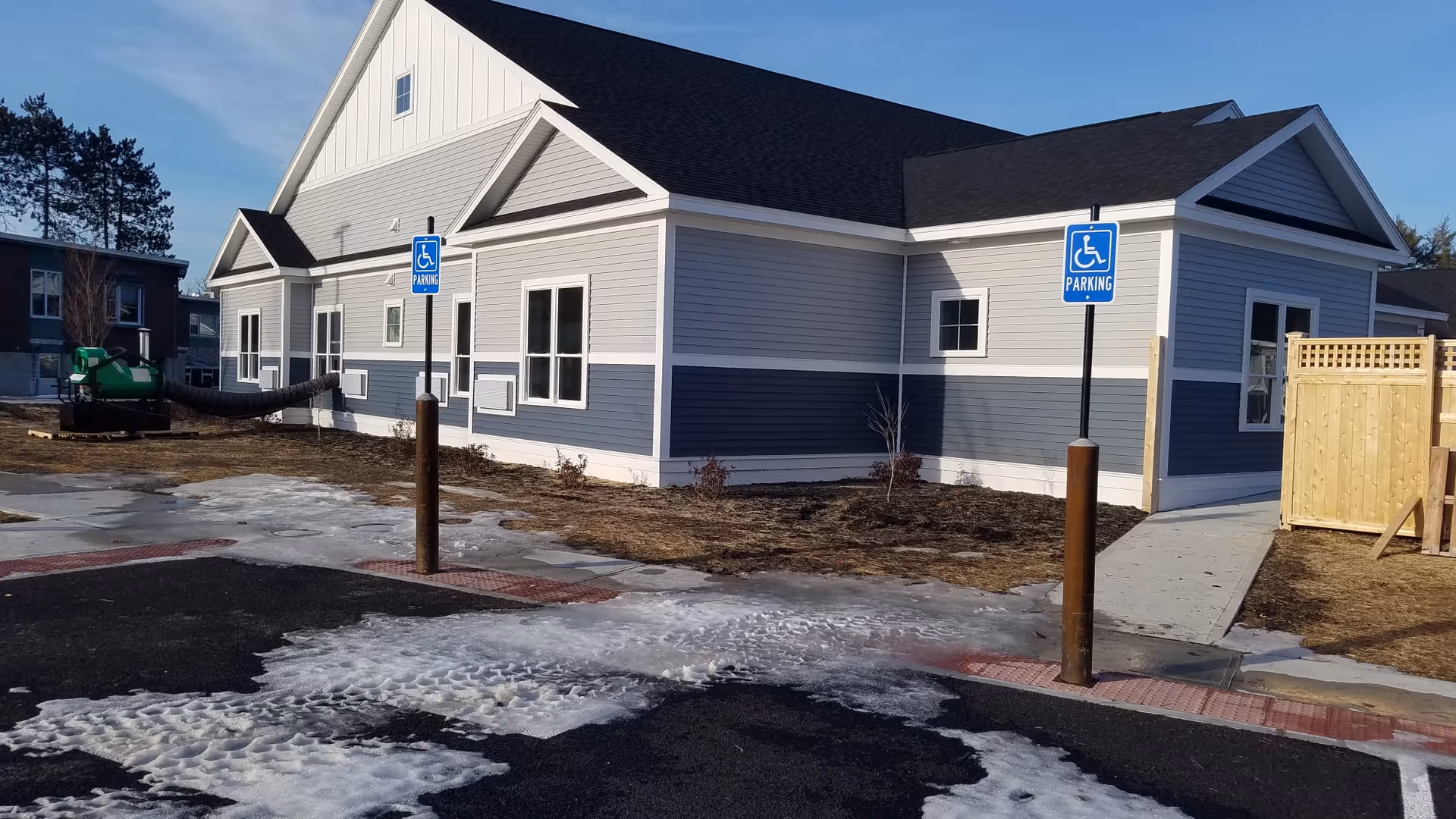 Exterior view of a single-story building with gray and white siding, a black roof, and multiple windows. There are two handicap parking signs in front of the building, a paved parking area with patches of snow and ice, and a wooden fence on the right side. The sky is clear and blue.