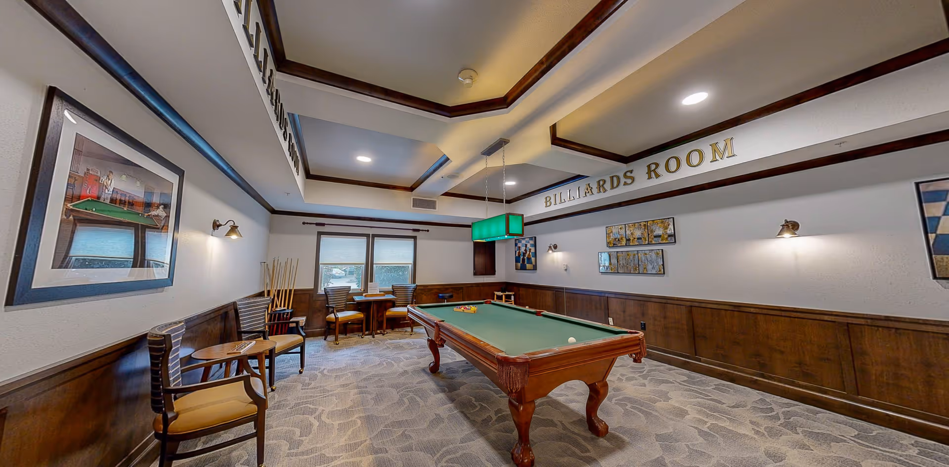 A billiards room with a pool table in the center, surrounded by chairs and small tables. The room has wood paneling on the lower walls, framed artwork, and a green hanging light fixture above the pool table. The ceiling has recessed lighting and decorative molding, with the words 'BILLIARDS ROOM' displayed prominently on the wall.