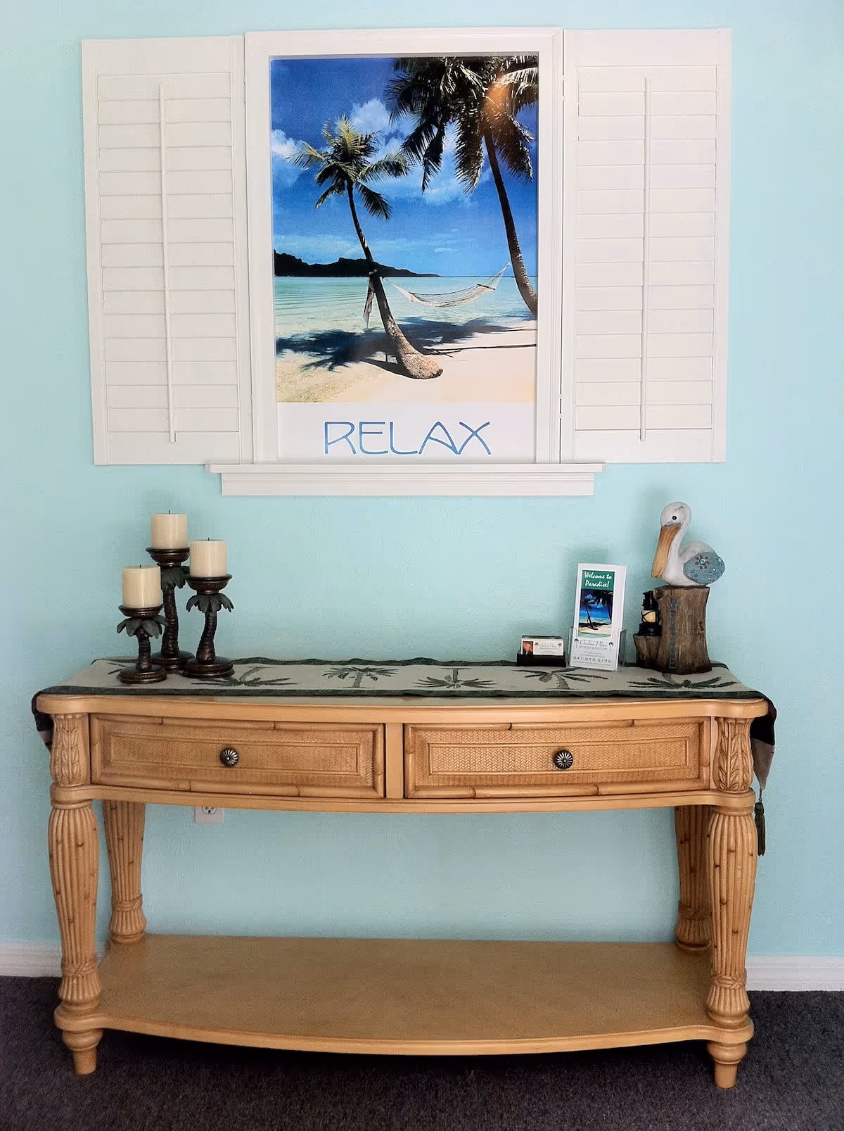 A wooden console table against a turquoise wall topped with candles, a pelican figurine, brochures and a framed beach poster reading 'RELAX'.