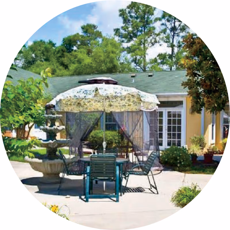 Outdoor patio area with a round table and several chairs under a floral-patterned umbrella with mesh curtains. There is a multi-tiered stone fountain to the left and a building with French doors and windows in the background, surrounded by green trees and shrubs.
