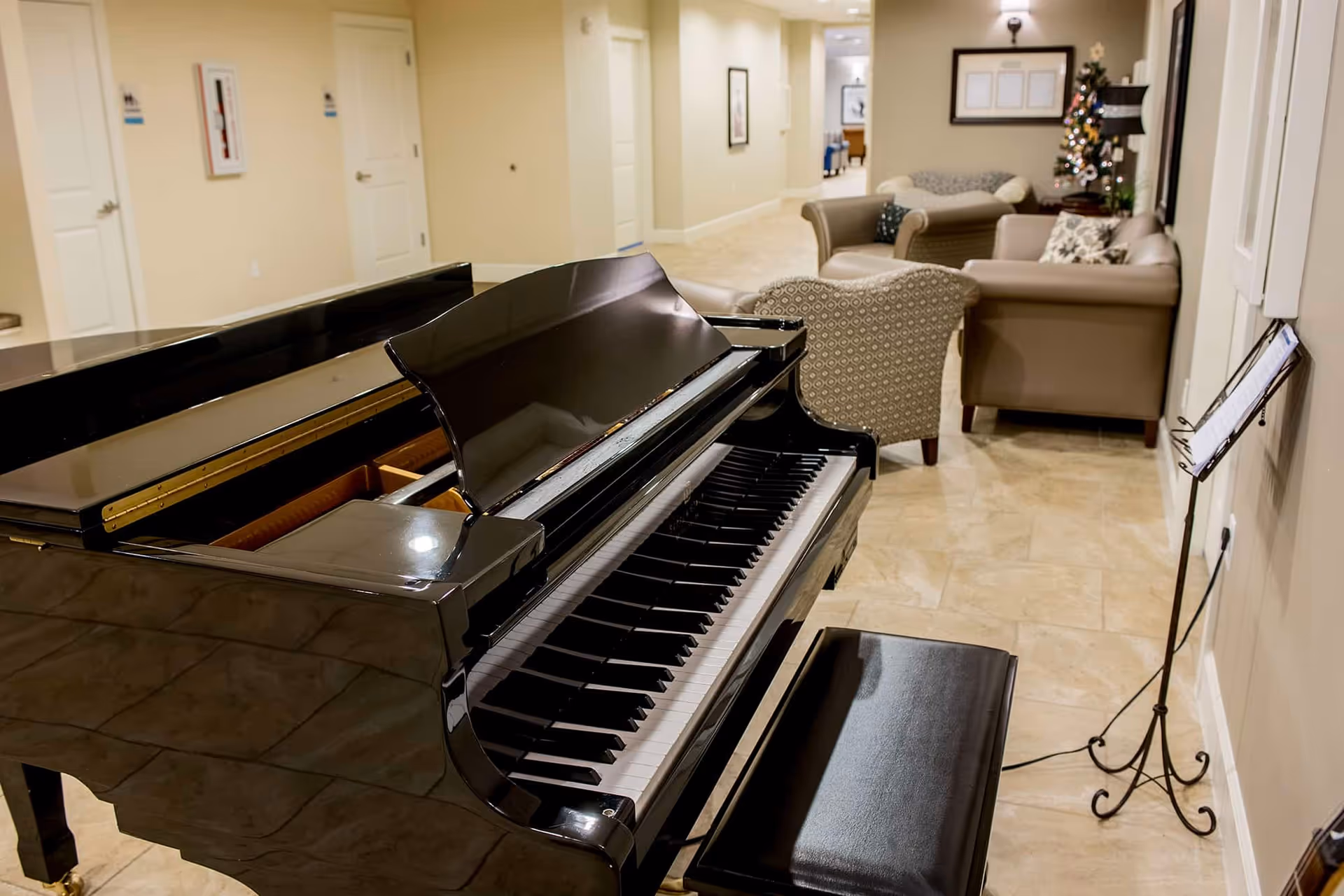 A black grand piano with a matching bench is positioned in a hallway area with beige tiled floors. In the background, there are several upholstered chairs and sofas arranged in a lounge setting. A small decorated Christmas tree is visible near the wall, along with framed pictures and a music stand holding sheet music.