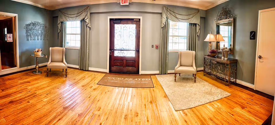 Wood‑floored assisted living entryway with a central decorative door flanked by windows, two upholstered chairs, and a console table with lamps.