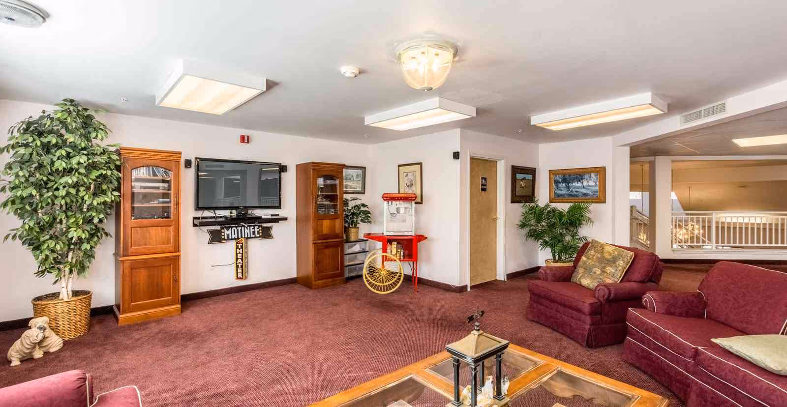 Communal lounge with burgundy sofas, a wall-mounted TV between wooden cabinets, a popcorn cart, plants and a central coffee table.